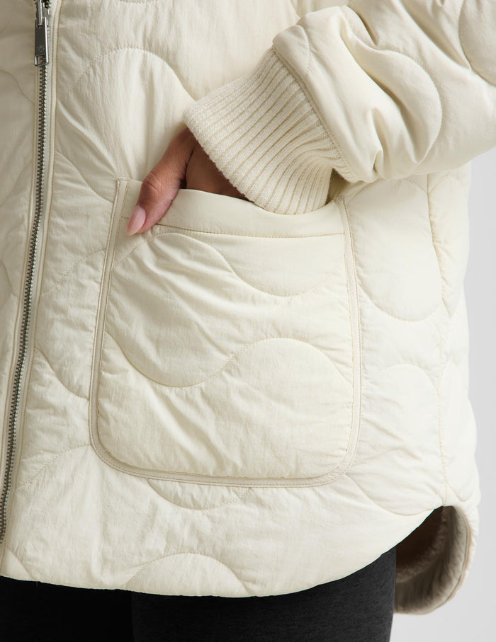 A woman modeling a cream-colored quilted zip-up jacket, paired with black leggings and silver sneakers, against a light gray studio background.