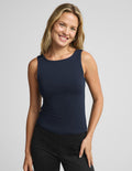 A smiling blonde woman wearing a fitted sleeveless navy tank top and black pants, photographed against a light gray studio background.