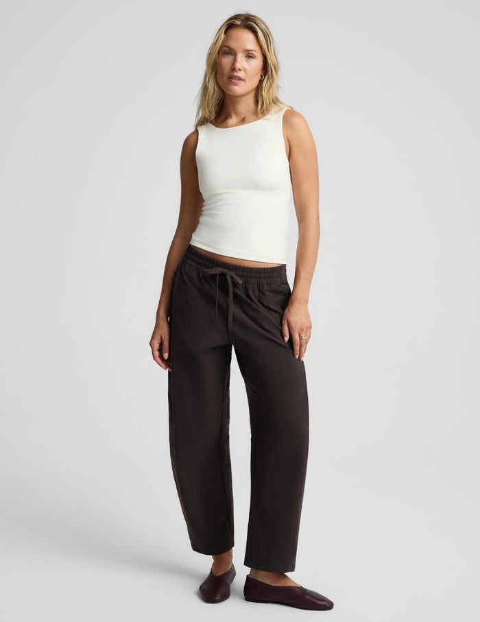 A blonde woman wearing a fitted sleeveless cream tank top and dark drawstring pants, photographed against a light gray studio background.