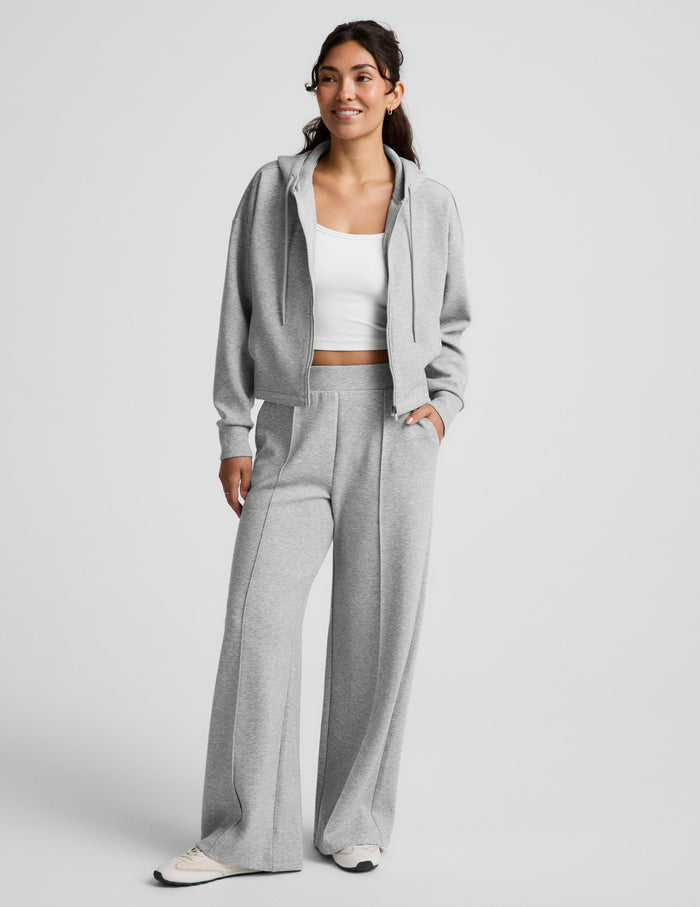 Woman wearing a gray zip-up sweatshirt and gray wide leg pants against a plain background.
