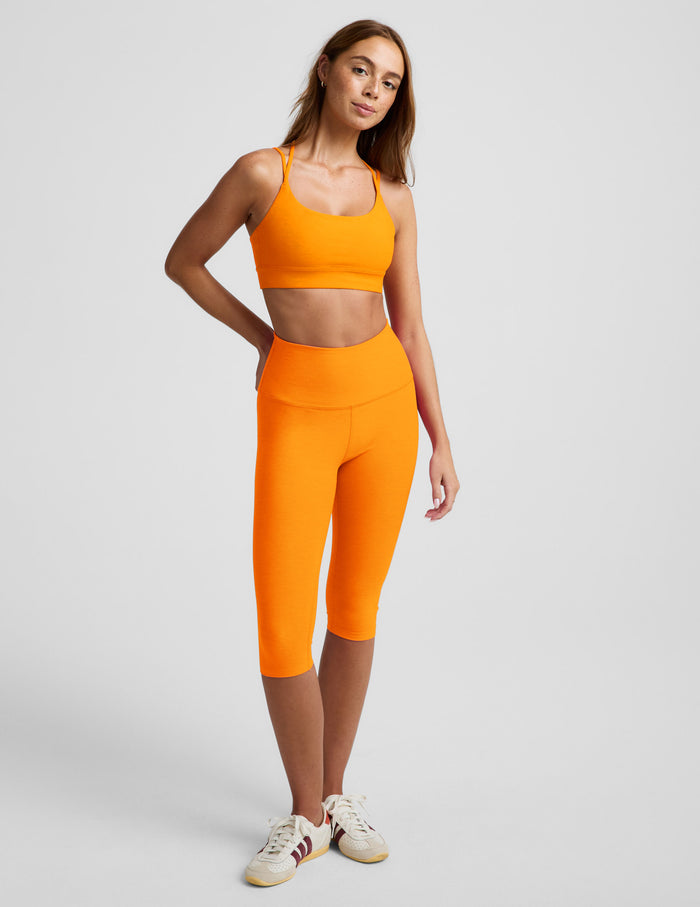 Woman wearing an orange strappy bra and orange leggings against a plain background.