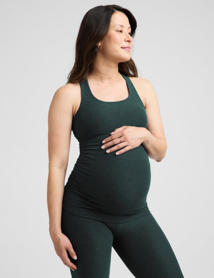 A pregnant woman wearing a dark forest green maternity tank top and matching leggings, standing against a light gray studio background.
