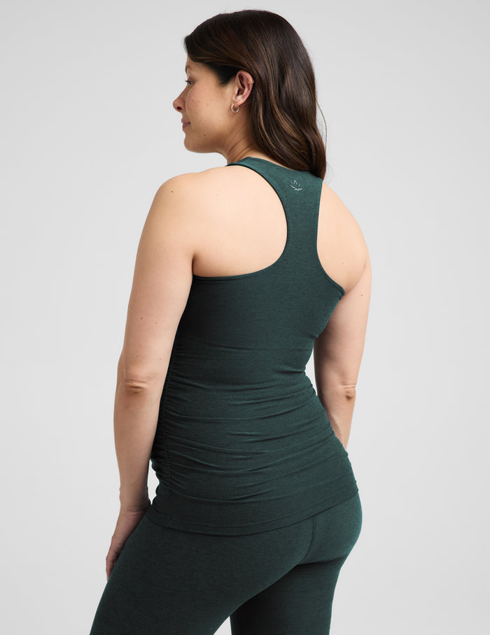 A pregnant woman wearing a dark forest green maternity tank top and matching leggings, standing against a light gray studio background.