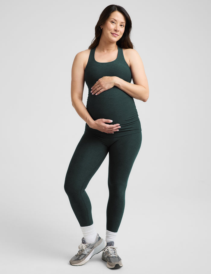 A pregnant woman wearing a dark forest green maternity tank top and matching leggings, standing against a light gray studio background.