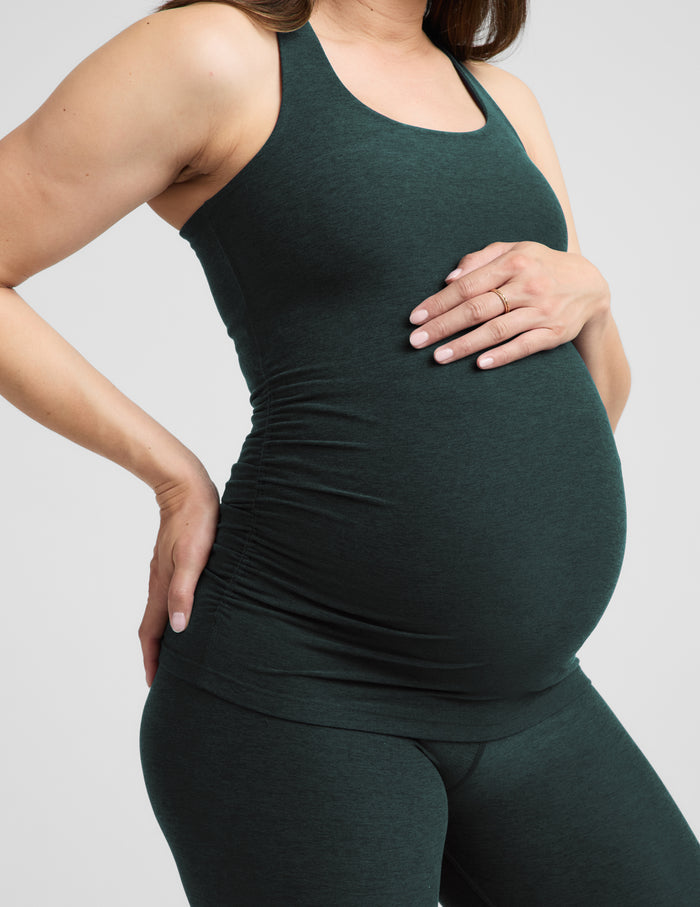 A pregnant woman wearing a dark forest green maternity tank top and matching leggings, standing against a light gray studio background.