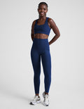 Woman wearing a blue sports bra and blue high-waisted midi leggings with pockets against a plain background.
