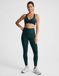 A woman modeling a matching dark green athletic set consisting of a sports bra and high-waisted leggings, paired with white sneakers, against a light gray studio background.