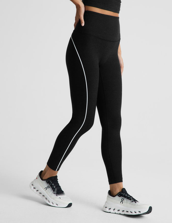 Black high-waisted athletic leggings with a white side stripe, modeled on a person wearing white and black running shoes against a light gray studio background.