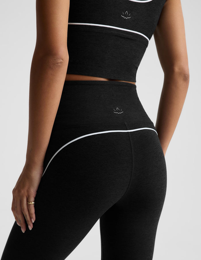 Close-up back view of a model wearing black athletic leggings and a matching crop top with thin white piping and a small white logo near the waistband.