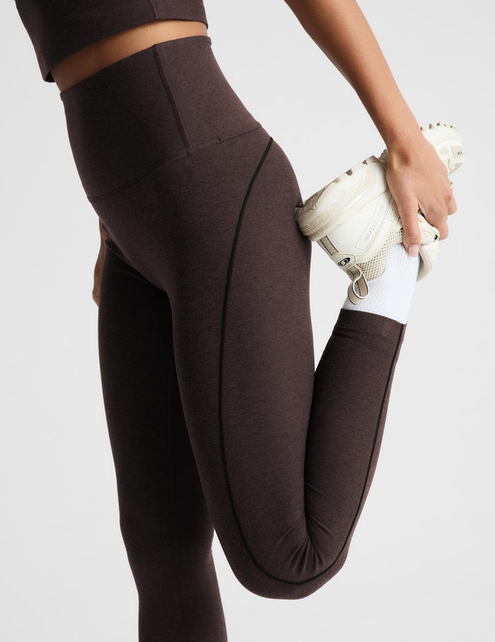 A close-up of a person wearing dark brown high-waisted leggings and a matching sports bra, stretching their leg while holding a white sneaker against a light gray studio background.