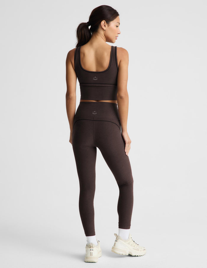 A woman wearing a matching dark brown activewear set consisting of a sports bra and high-waisted leggings, shown from a rear three-quarter angle against a light gray studio background.