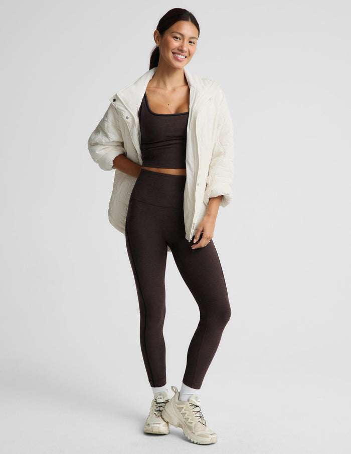 A woman modeling a dark brown activewear set consisting of a sports bra and high-waisted leggings, styled with an open cream-colored lightweight jacket and beige sneakers against a light gray studio background.