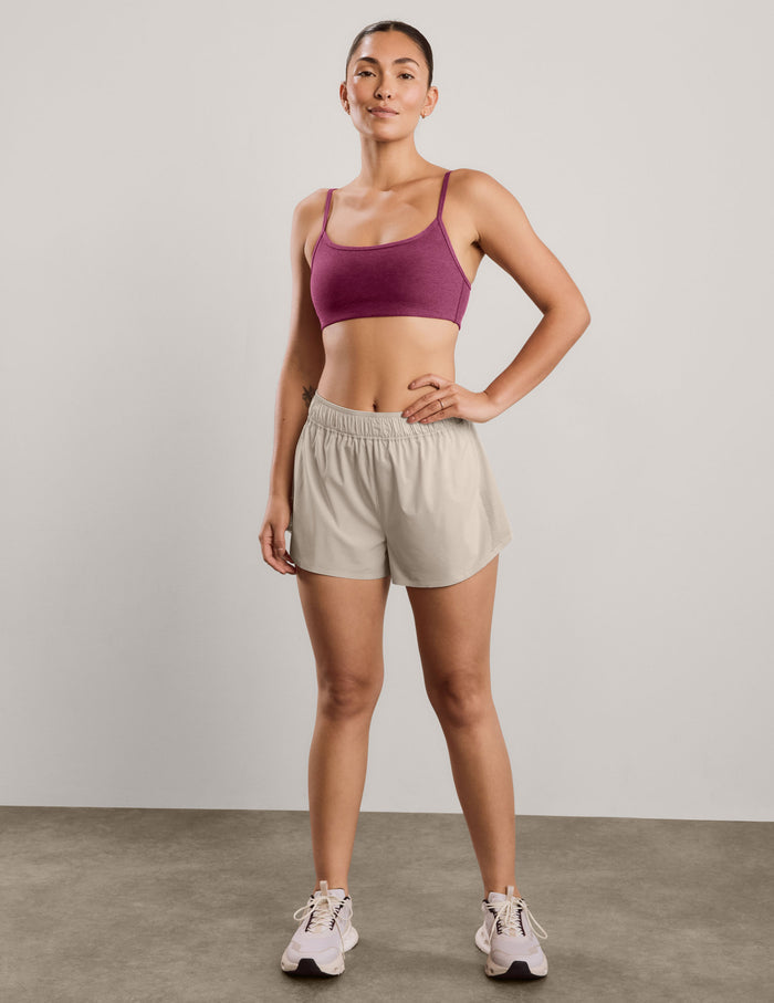 A woman modeling a heathered red purple sports bra and light beige athletic shorts against a solid light gray studio background.