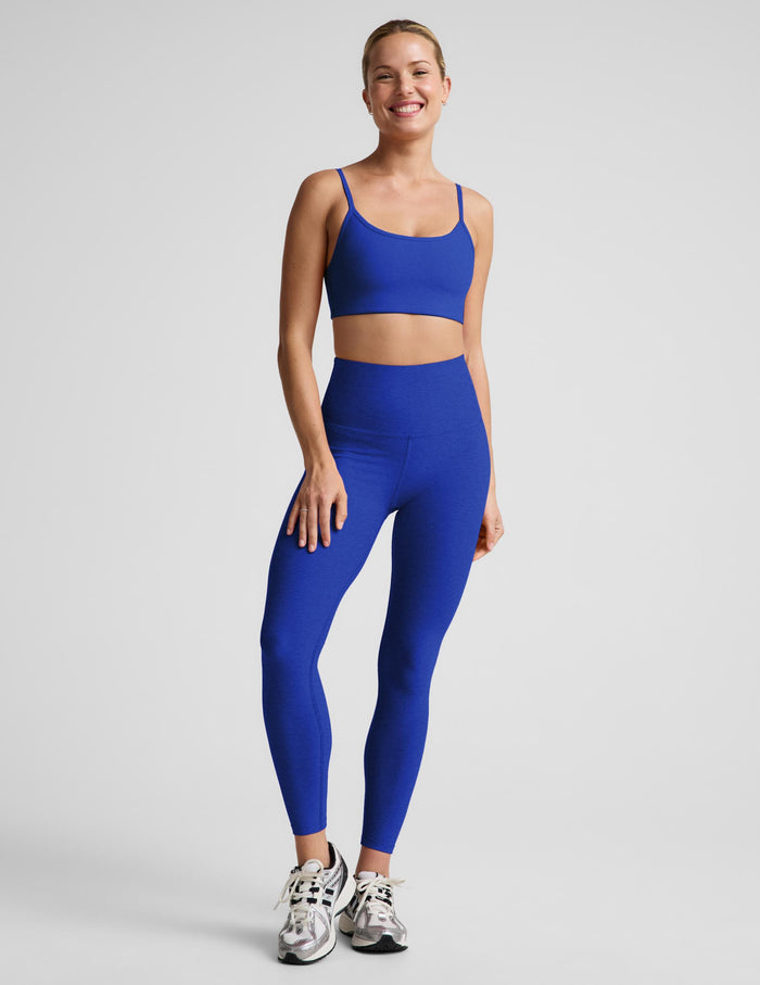A woman modeling a matching royal blue activewear set consisting of a sports bra and high-waisted leggings, paired with white and gray sneakers, against a light gray studio background.