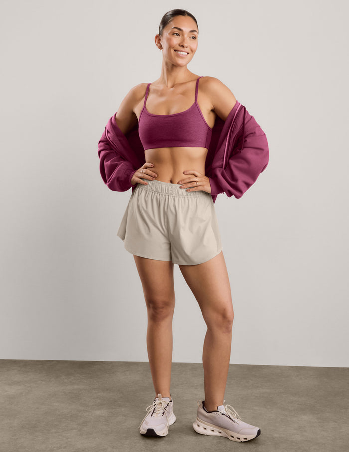 A woman modeling a heathered red purple sports bra and light beige athletic shorts against a solid light gray studio background.