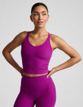 A woman modeling a matching athletic set consisting of a scoop-neck crop top and high-waisted biker shorts in a vibrant magenta-purple color, set against a plain light gray studio background.