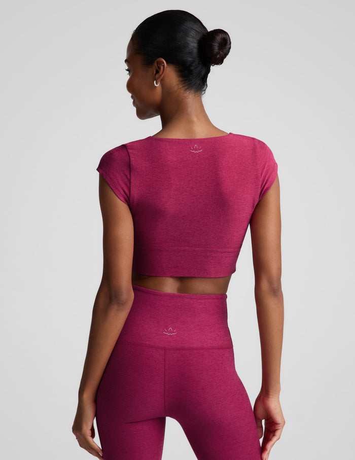 A woman modeling a matching athletic set featuring a short-sleeve crop top with a twisted front detail and high-waisted leggings, in a heathered magenta-pink color, against a solid light gray background.