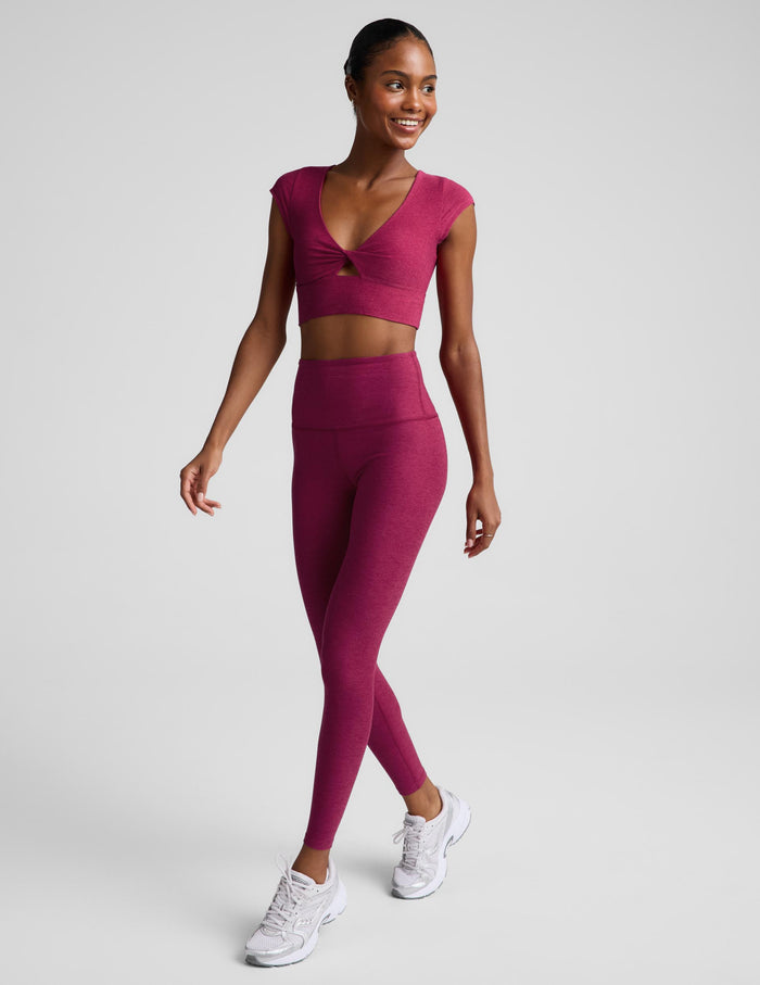A woman modeling a matching athletic set featuring a short-sleeve crop top with a twisted front detail and high-waisted leggings, in a heathered magenta-pink color, against a solid light gray background.