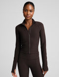 A woman modeling a dark brown long-sleeve athletic zip-up jacket with a stand collar and matching dark brown leggings against a light gray studio background.