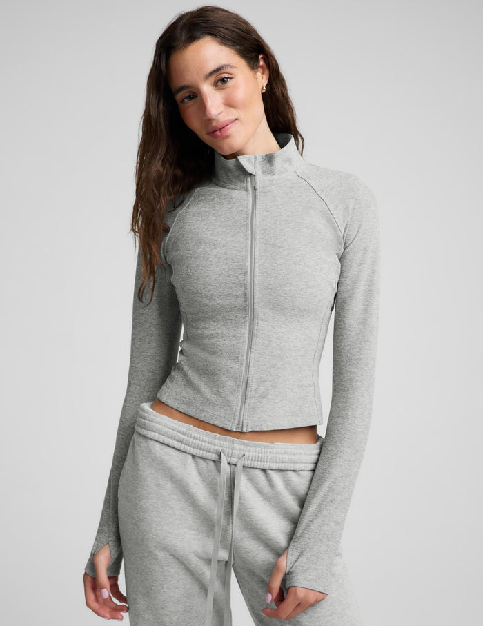 A woman modeling a light gray heather cropped zip-up athletic jacket with matching jogger sweatpants against a light gray studio background.