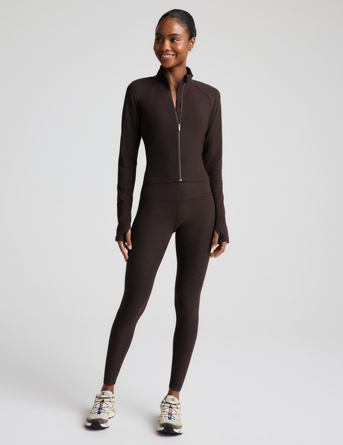 A woman modeling a dark brown jacket and matching high-waisted leggings, paired with white sneakers, against a light gray studio background.