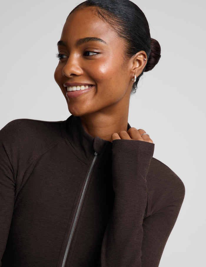 A smiling woman with her hair in a bun wearing a dark brown long-sleeve zip-up athletic top, shown in a close-up profile shot against a light gray background.