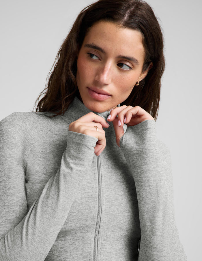 A woman modeling a light gray heather cropped zip-up athletic jacket with matching jogger sweatpants against a light gray studio background.