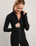 Woman wearing a black full-length zip-up jacket and black biker shorts against a plain background.