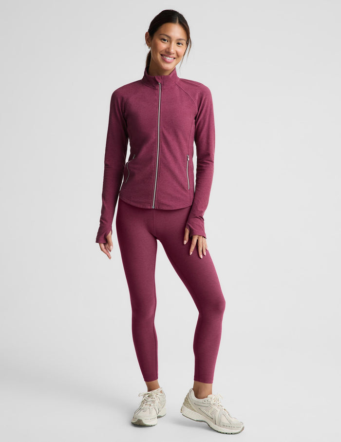 A woman modeling a matching burgundy athletic set consisting of a long-sleeve zip-up jacket and high-waisted leggings, paired with off-white sneakers, against a light gray studio background.