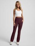 A woman modeling a pair of high-waisted, straight-leg athletic pants in heathered burgundy, paired with a white crop top and white sneakers, against a light gray studio background.