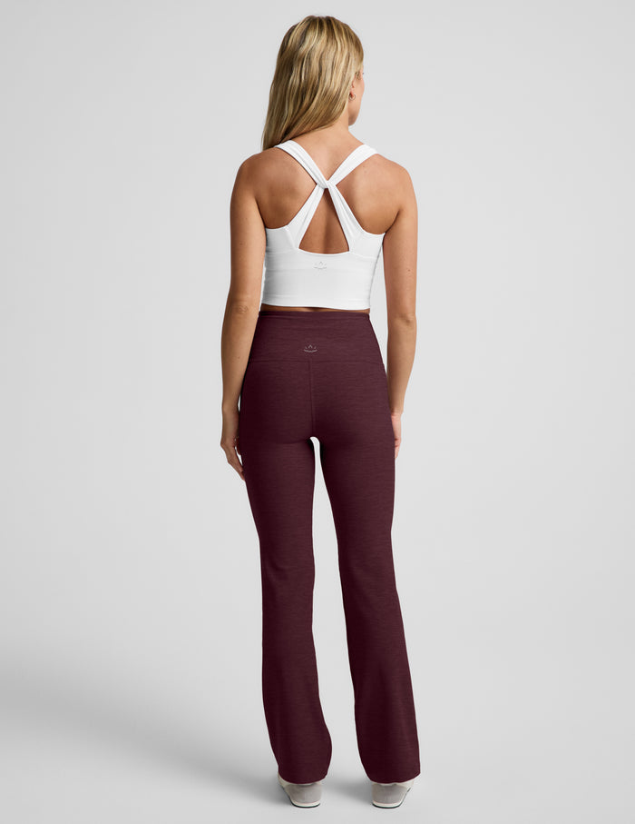 A woman modeling a pair of high-waisted, straight-leg athletic pants in heathered burgundy, paired with a white crop top and white sneakers, against a light gray studio background.