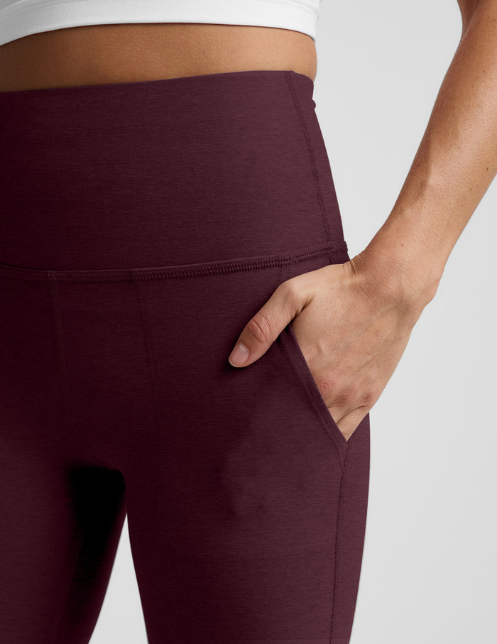 Close-up of a model wearing high-waisted burgundy leggings with a side pocket, shown from the waist down against a light gray background.