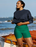 A woman standing on a beach next to a wooden boat, wearing a dark navy long-sleeve crewneck sweatshirt and bright green high-waisted biker shorts.