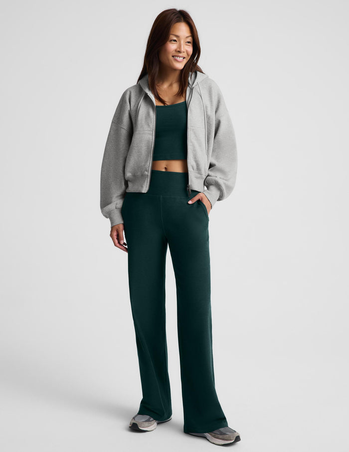 A woman wearing a gray zip-up hooded jacket, dark green racerback cropped tank top and dark green wide leg pants against a plain studio background.