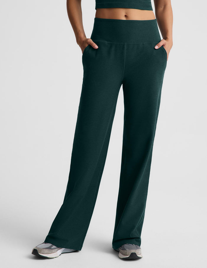 A woman wearing a dark green racerback cropped tank top and dark green wide leg pants against a plain studio background.