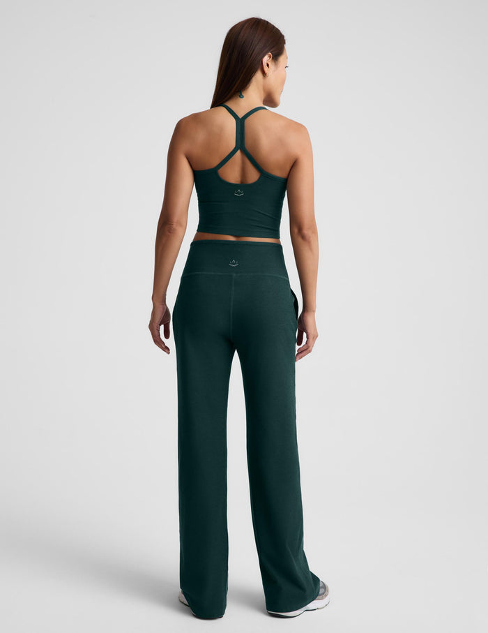 A woman wearing a dark green racerback cropped tank top and dark green wide leg pants against a plain studio background.