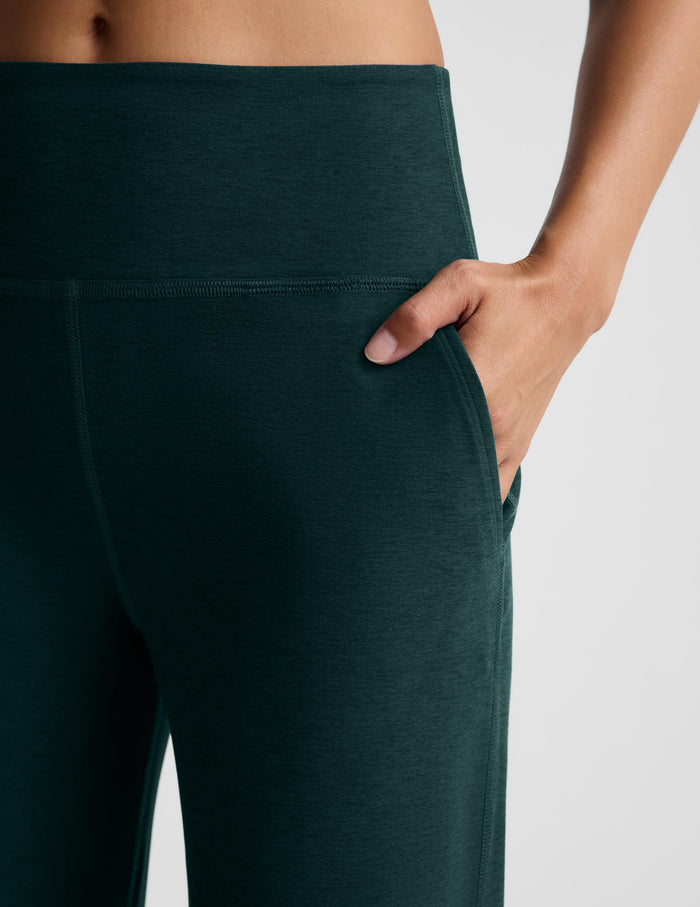 Close-up of a hand in the pocket of dark green wide leg pants against a plain studio background.