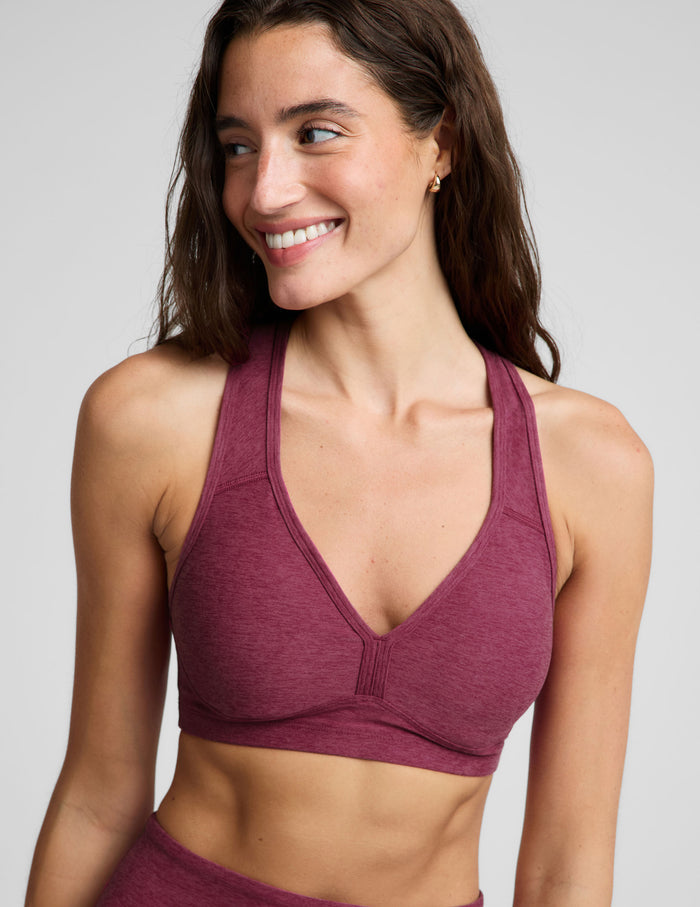 A woman modeling a heathered burgundy sports bra and matching high-waisted pants against a light gray studio background.