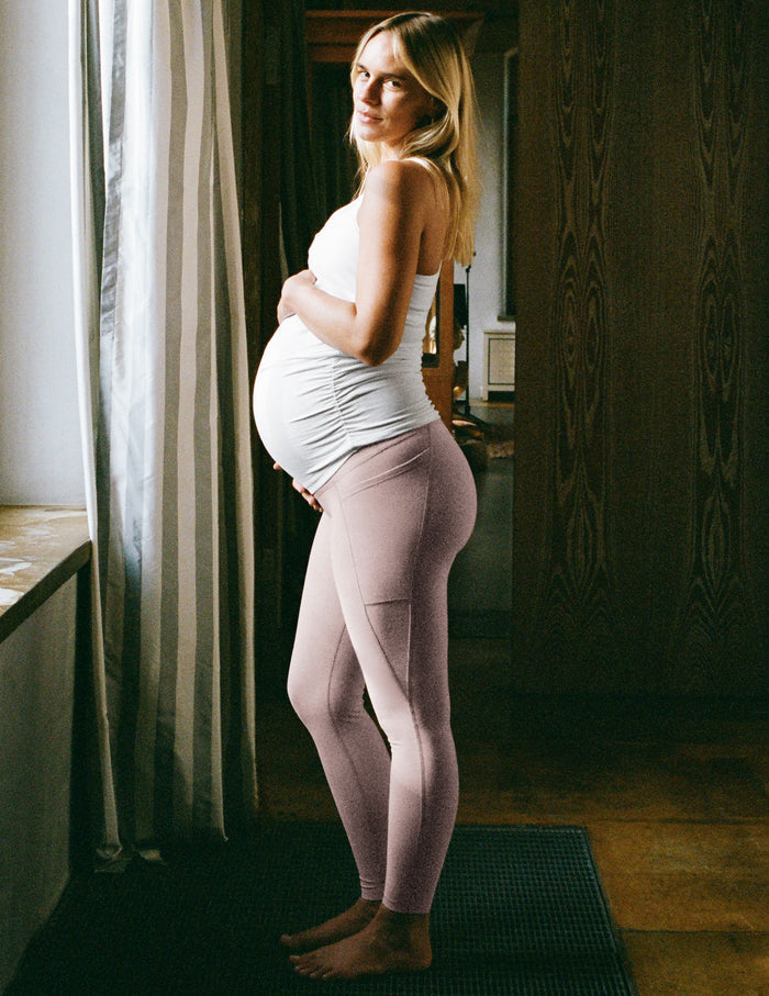 Woman wearing a white maternity tank top and pink maternity leggings with pockets.