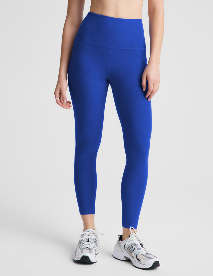 A woman modeling high-waisted royal blue leggings with a white sports bra and white sneakers, standing against a light gray studio background.