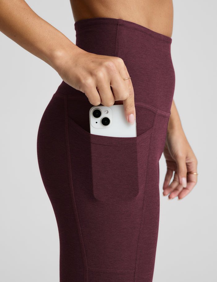 Close-up of a person wearing heathered burgundy high-waisted leggings with a side pocket, shown holding a white smartphone in the pocket against a light gray studio background.