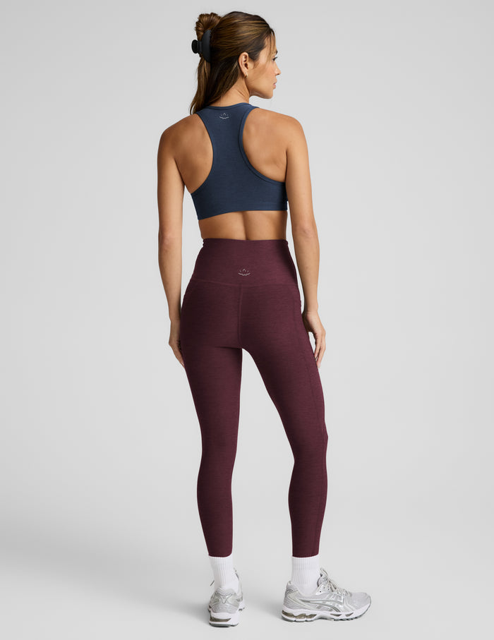 A woman shown from the back wearing a navy blue sports bra and high-waisted burgundy leggings, standing against a light gray studio background.