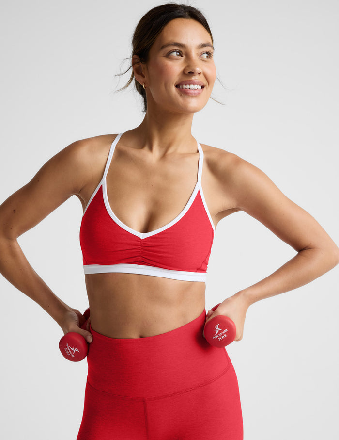 Woman in red sports bra and leggings holding red dumbbells against a light gray background.