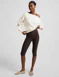 Woman wearing a brown bra, pedal pusher leggings, and a white off the shoulder fleece sweatshirt against a plain background.