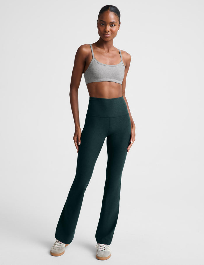 A model wearing dark forest green high-waisted bootcut leggings and a light gray sports bra, standing against a light gray studio background.