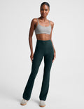 A model wearing dark forest green high-waisted bootcut leggings and a light gray sports bra, standing against a light gray studio background.