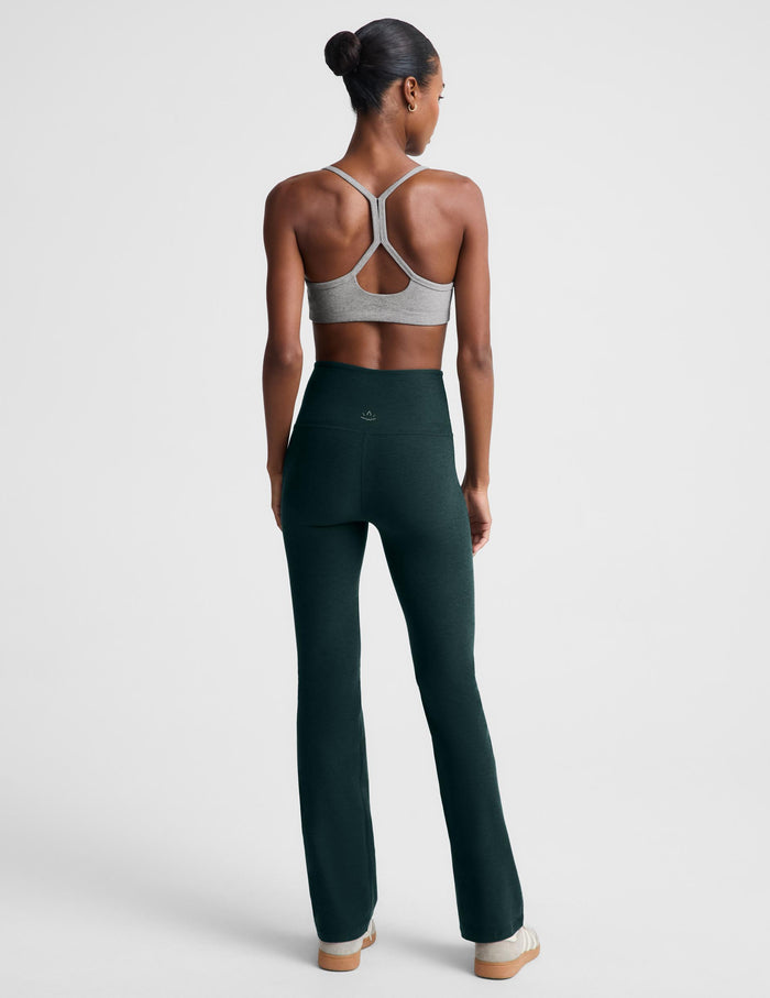 A model wearing dark forest green high-waisted bootcut leggings and a light gray sports bra, standing against a light gray studio background.
