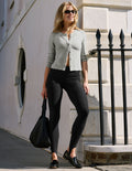 A woman stands outdoors on a sidewalk wearing a light gray cropped cardigan, black leggings, black loafers, and sunglasses, holding a black woven handbag. She is posed in front of a pale stone building with a black metal fence.