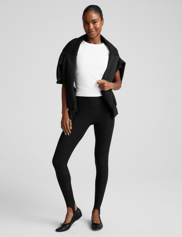 A woman modeling black leggings, a white top, and a black draped jacket, standing against a light gray studio background.