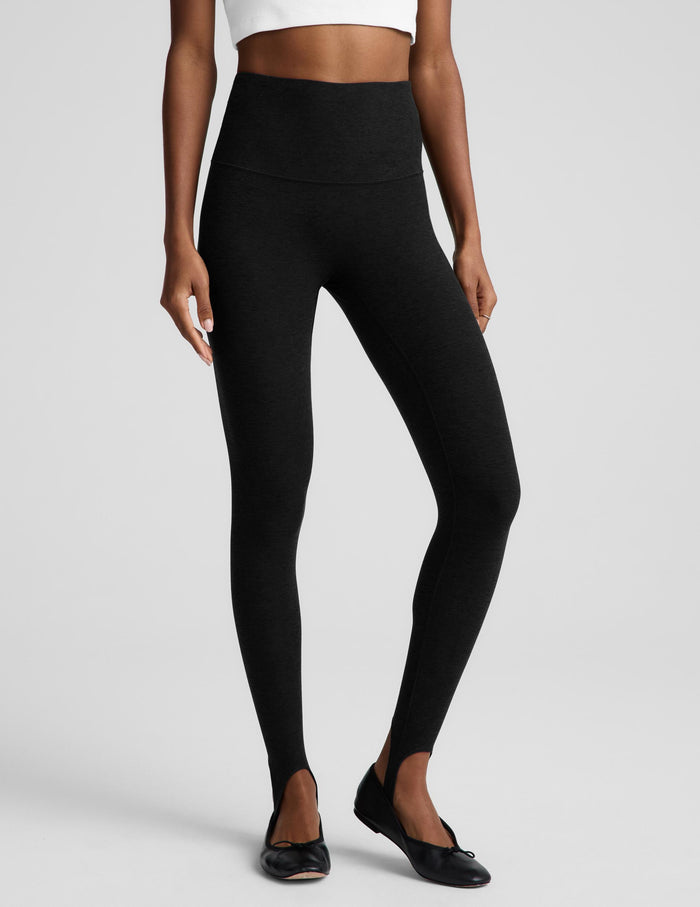 A woman modeling black leggings, a white top, and a black draped jacket, standing against a light gray studio background.